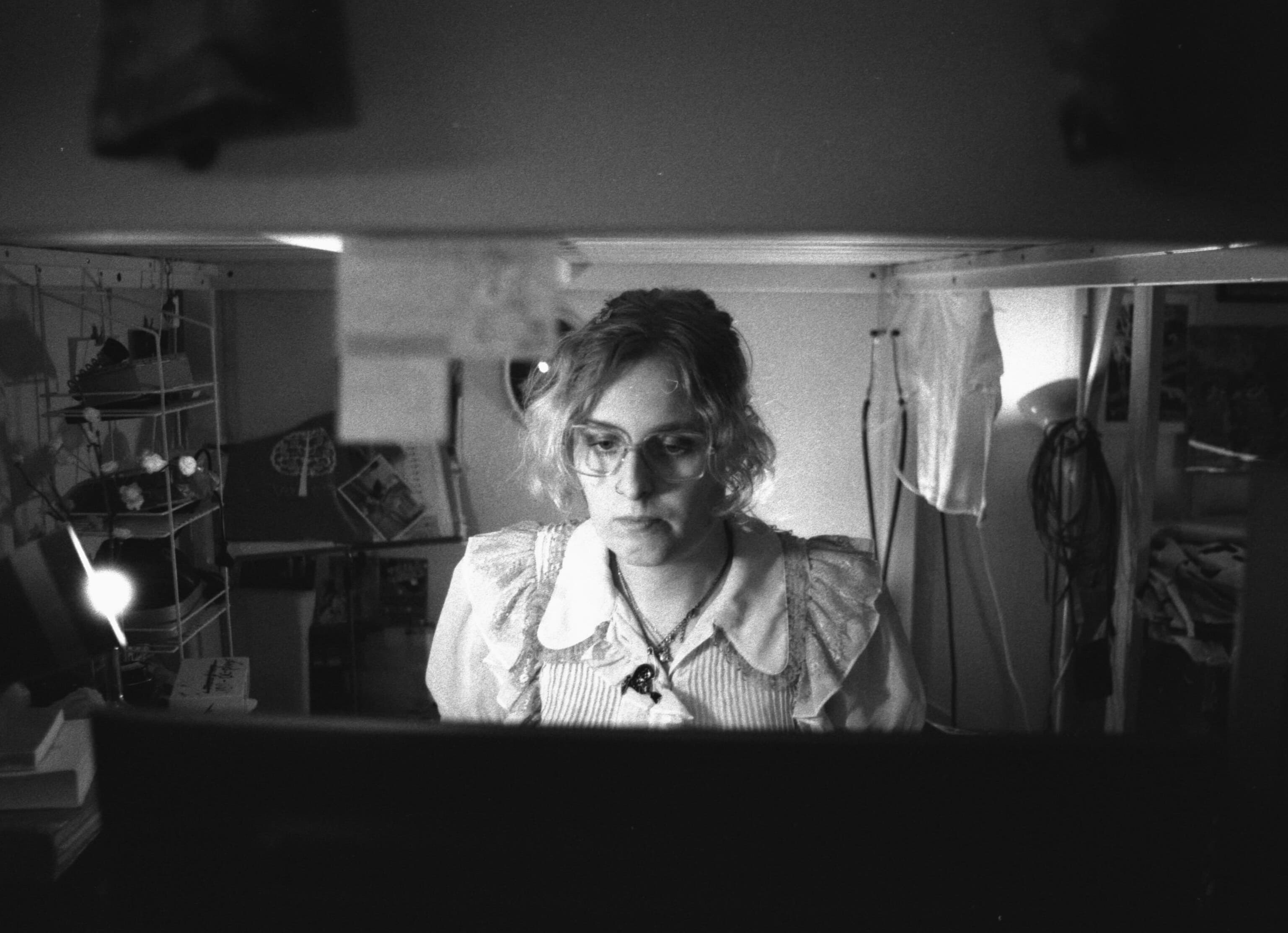 A person with glasses and curly hair is focused on a screen in a dimly lit, cluttered room.